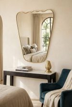 Handmade wavy top brass wall mirror with tapered sides and solid brass frame displayed above bedroom console.